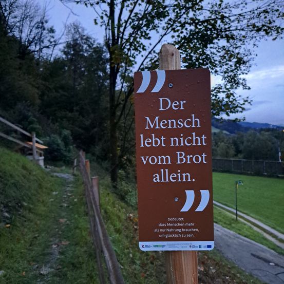 Ein Schild mit einem deutschen Zitat, das besagt 'Der Mensch lebt nicht vom Brot allein', befindet sich auf einem Holzpfahl. Es steht auf einem Rasen mit einem Pfad und Bäumen.