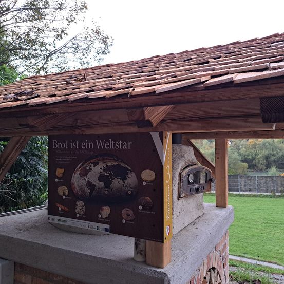 Ein hölzernes Pavillon mit einem Ziegeldach schützt einen Pizzaofen. Ein Informationsschild mit dem Titel 'Brot ist ein Weltstar' zeigt verschiedene Brotsorten. Im Hintergrund befindet sich ein eingezäuntes Feld mit Grünpflanzen.