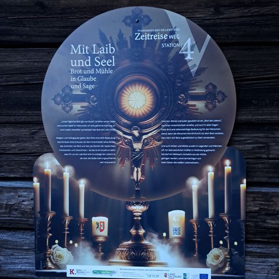 Ein kreisförmiges Schild zeigt eine religiöse Szene mit Kerzen, einem Kelch und einem Kreuz. Es steht 'Mit Leib und Seel' und 'Brot und Mühle in Glaube und Sage', wahrscheinlich ein deutscher Ausdruck über Körper, Seele, Brot und Mühle im Glauben und in der Sage. Der Hintergrund ist dunkel mit einer Holzstruktur.