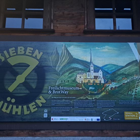 Ein großes Schild an einer Holzwand mit einer Zeichnung einer Kirche und Bergen, das das Siebenbuehllen-Logo und Text über das Freilichtmuseum & Brot Way zeigt.