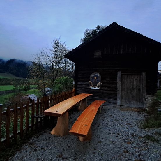 Ein rustikales Holzhaus mit einer Holztür steht neben zwei Holzbänken auf einem Schotterbereich. Es hat einen Holzzaun davor und ein rundes Fenster an der Wand.