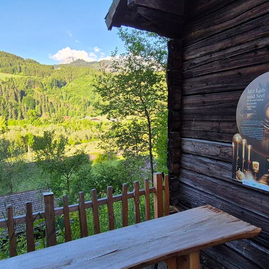 Eine Holzbank steht vor einer Holzhütte mit Blick auf Berge und Bäume. An der Wand der Hütte befindet sich ein Schild.
