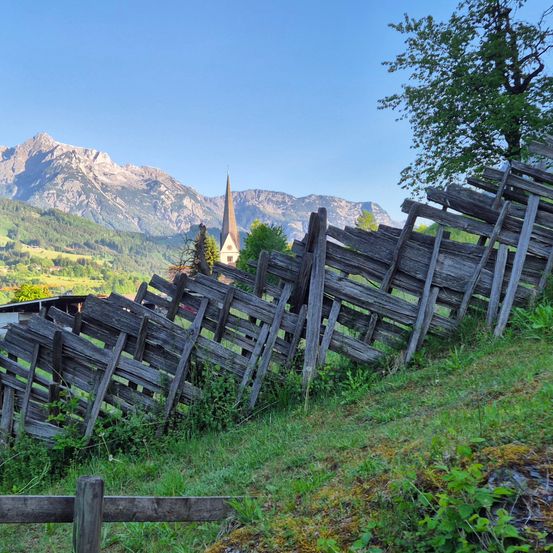 Ein hölzerner Zaun am Hang neigt sich zu einer Kirche mit einem Kirchturm unter einem blauen Himmel mit Bergen im Hintergrund.