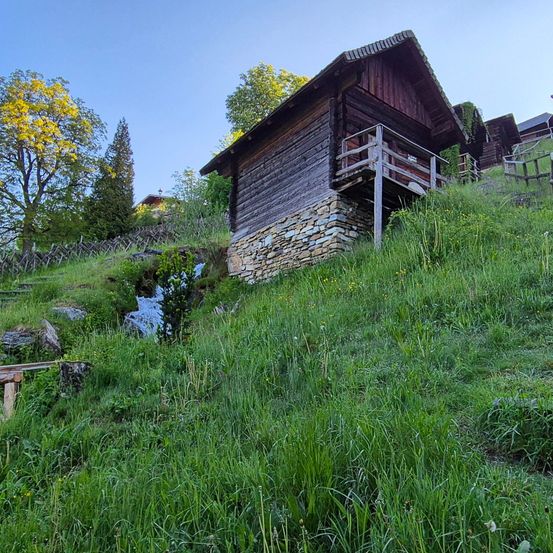 Ein rustikales Holzhaus steht auf einem grün bewachsenen Hang mit einem kleinen Bach in der Nähe. Das Haus hat ein Steinfundament und eine Holzveranda. Im Hintergrund sind Bäume und Sträucher zu sehen.