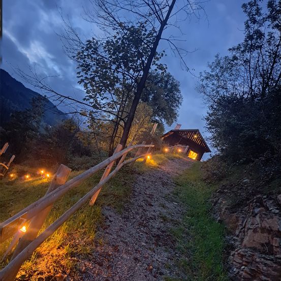 Ein kleines Holzhaus am oberen Rand eines Hügels mit brennenden Kerzen auf einem Holzzaun. Das Haus ist von Bäumen und Bergen umgeben.