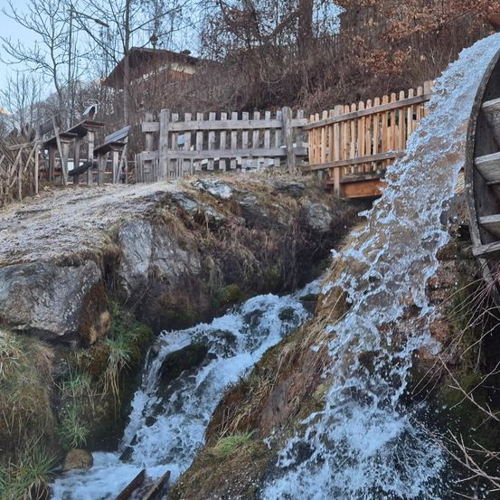 Ein Wasserfall stürzt an Felsen vorbei, nahe einem Holzzaun, umgeben von Bäumen mit braunen Blättern. Hinter dem Zaun befindet sich eine Holzkonstruktion und ein Berg in der Ferne.