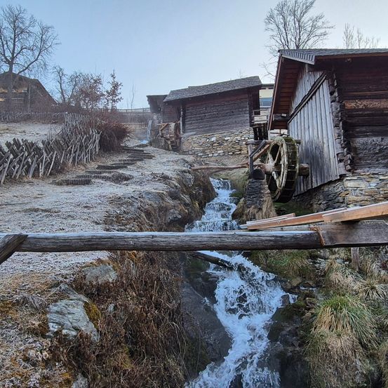 Eine Winterszene mit einer kleinen Holzbrücke über einem Bach. Wasser fließt über Steine in der Nähe alter Stein- und Holzgebäude. Bäume ohne Blätter umgeben das Gebiet.