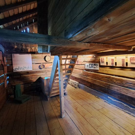 Ein Innenraum einer Holzhütte mit Holzboden, Holzwänden und einer Holztreppe. Ein grüner Stuhl befindet sich auf der linken Seite und ein Holzschrank auf der rechten. Es hängen Holzfässer an der Wand und einige Rahmen sind an der Wand montiert.