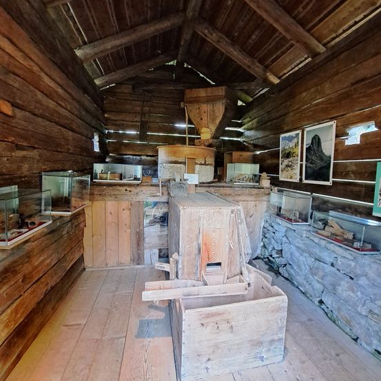 Ein hölzerner Raum mit Holzböden und -wänden, mehreren Glasvitrinen und einem großen Holzobjekt in der Mitte. Ein Foto ist an der Wand.