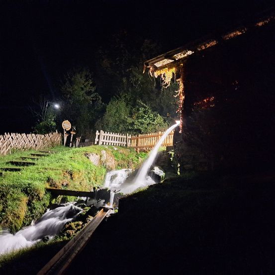 Eine nächtliche Ansicht eines kleinen Wasserfalls, der aus einem Gebäude in einen Rasenbereich fließt, beleuchtet von einer Straßenlampe und einem Licht aus dem Gebäudeinneren.