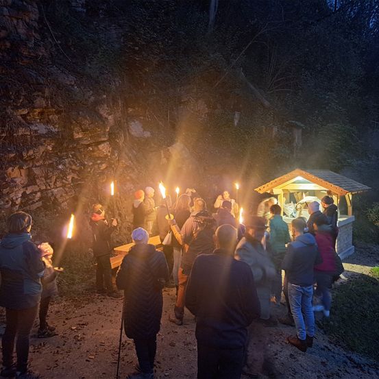 Eine Gruppe von Menschen mit brennenden Fackeln steht vor einem kleinen Gebäude im Wald. Das Gebäude ist beleuchtet, und die Menschen tragen Jacken und Mützen. Das Gebiet ist von Bäumen und Felsen umgeben.
