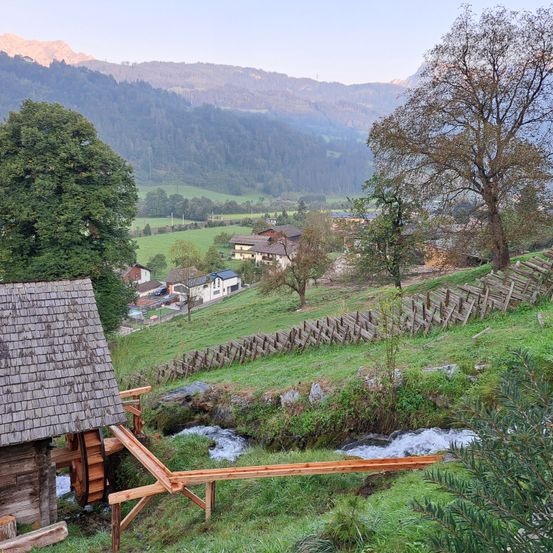 Ein rustikales Holzhaus steht auf einem Hügel mit einem fließenden Bach. Das Haus hat ein Holzrad in der Nähe des Wassers. Im Hintergrund befinden sich Häuser, ein Feld und Berge.