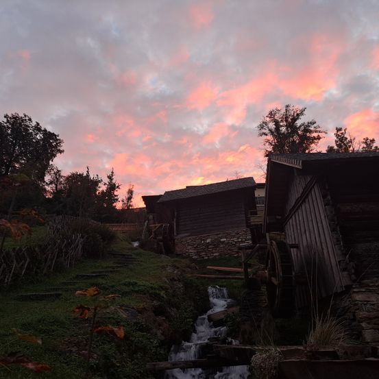 Eine malerische Dorfszene bei Sonnenuntergang mit alten Holzgebäuden und einem Wasserrad. Der Himmel ist in Rosa- und Orangetöne getaucht.