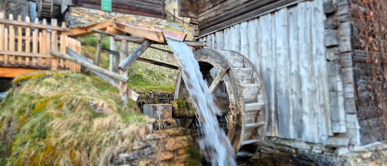 Ein altes Holzrad steht in der Nähe eines Gebäudes. Wasser fließt durch eine Holzrinne in das Rad.
