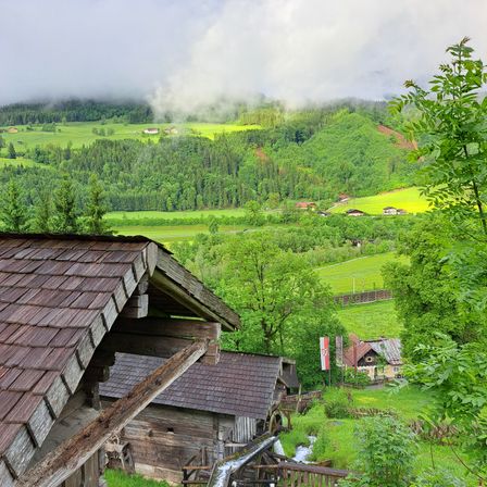 Eine ländliche Szene mit einem Holzhaus, einem Bach und üppigen grünen Hügeln. Das Wetter ist bewölkt mit Nebel in der Ferne.
