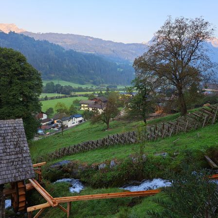 Ein malerisches Alpental mit einem Holzzaun und einem Wasserrad. Bergrücken und grüne Wiesen im Hintergrund.