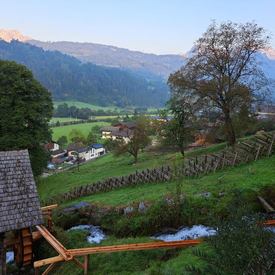 Ein malerisches Alpental mit einem Holzzaun und einem Wasserrad. Bergrücken und grüne Wiesen im Hintergrund.
