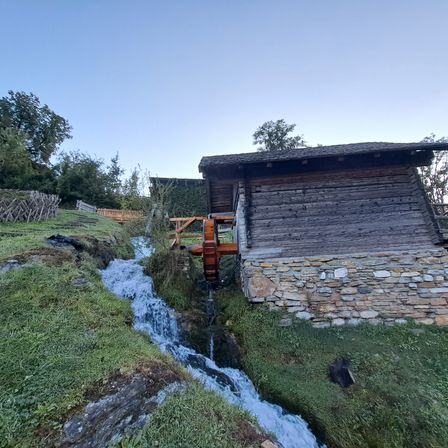 Ein altes Holzgebäude mit Steinfundament steht an einem Hang mit fließendem Wasser. Bäume umgeben das Gebiet.