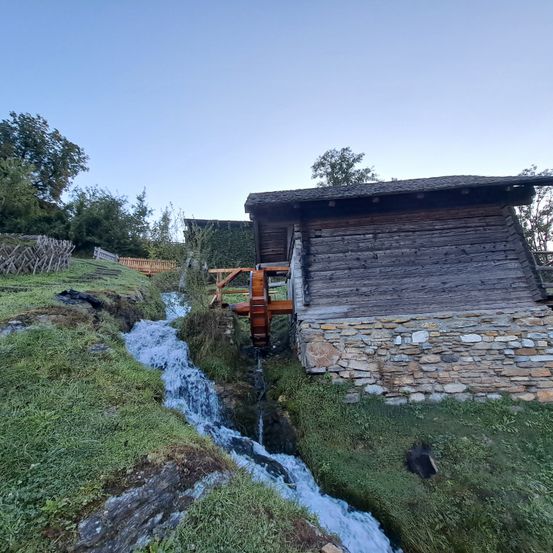 Ein altes Holzgebäude mit Steinfundament steht an einem Hang mit fließendem Wasser. Bäume umgeben das Gebiet.