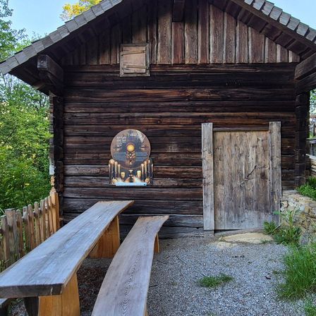 Eine rustikale Holzhütte mit einer Holztür und einer kleinen Tafel mit Kerzen an der Wand. Zwei Holzbänke stehen draußen.