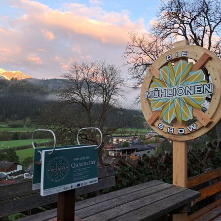 Eine landschaftliche Ansicht eines Berggebiets mit einem 'Die Muhlionen Show'-Schild, einem Quizmaster-Brett und einer Holzbank im Vordergrund.