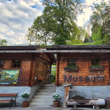 Ein kleines Holzgebäude mit der Aufschrift Museum, Blumenkästen an den Fenstern und einem großen Rad dahinter. Bäume umgeben das Gebiet, und es gibt Stufen, die zum Eingang führen.