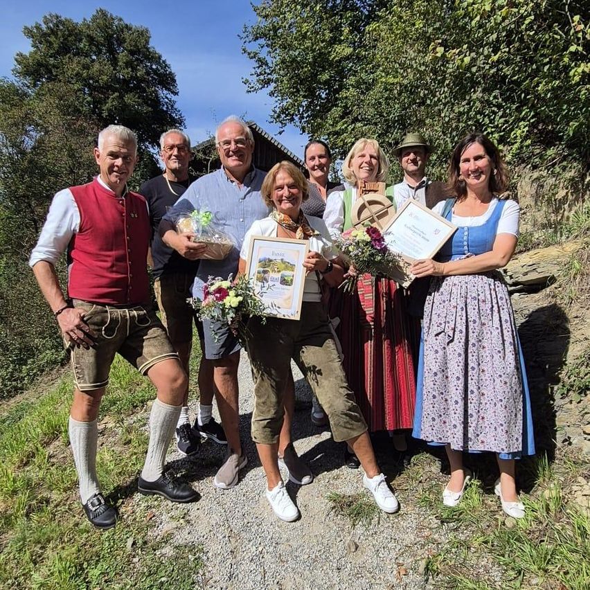 Eine Gruppe von Menschen in traditioneller bayerischer Kleidung posiert für ein Foto auf einem Kiesweg, einige halten Blumen und Urkunden.