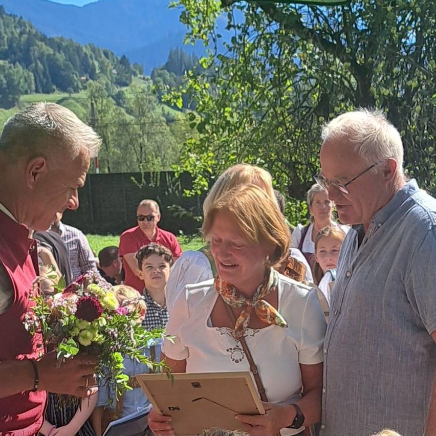 Ein Paar steht lächelnd vor einer Menschenmenge, wobei einer der Männer einen Blumenstrauß und der andere ein gerahmtes Dokument hält. Sie sind von einer Gruppe von Menschen umgeben, mit einem malerischen Berghintergrund.