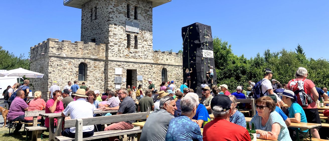 Eine Gruppe von Menschen versammelt sich um Tische vor einer steinernen Burg. Einige Personen klettern an einer an der Burg befestigten Kletterwand. Ein klarer blauer Himmel ist über ihnen.