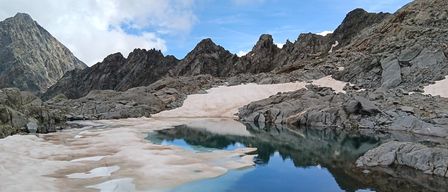 Eine hochgelegene Berglandschaft mit felsigem Gelände und einem kleinen, klaren See, der den blauen Himmel widerspiegelt. Schneeflecken zieren die felsige Oberfläche.