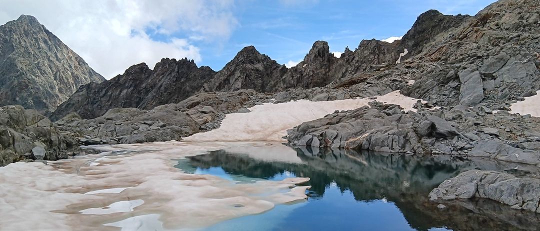 Eine hochgelegene Berglandschaft mit felsigem Gelände und einem kleinen, klaren See, der den blauen Himmel widerspiegelt. Schneeflecken zieren die felsige Oberfläche.