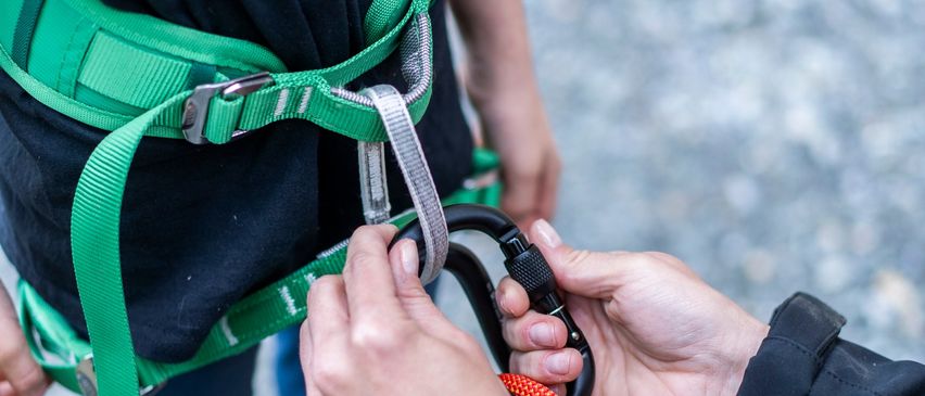 Eine Person justiert einen Karabiner an einem Gurtzeug, während eine andere Person dahinter steht. Das Gurtzeug ist grün und grau, und der Karabiner ist schwarz.