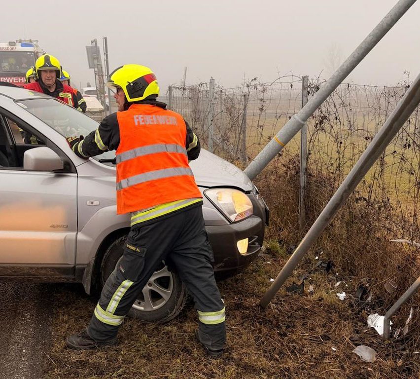 Two firefighters in orange vests assist at a car accident site, one holding the car's door, and another walking beside the car.