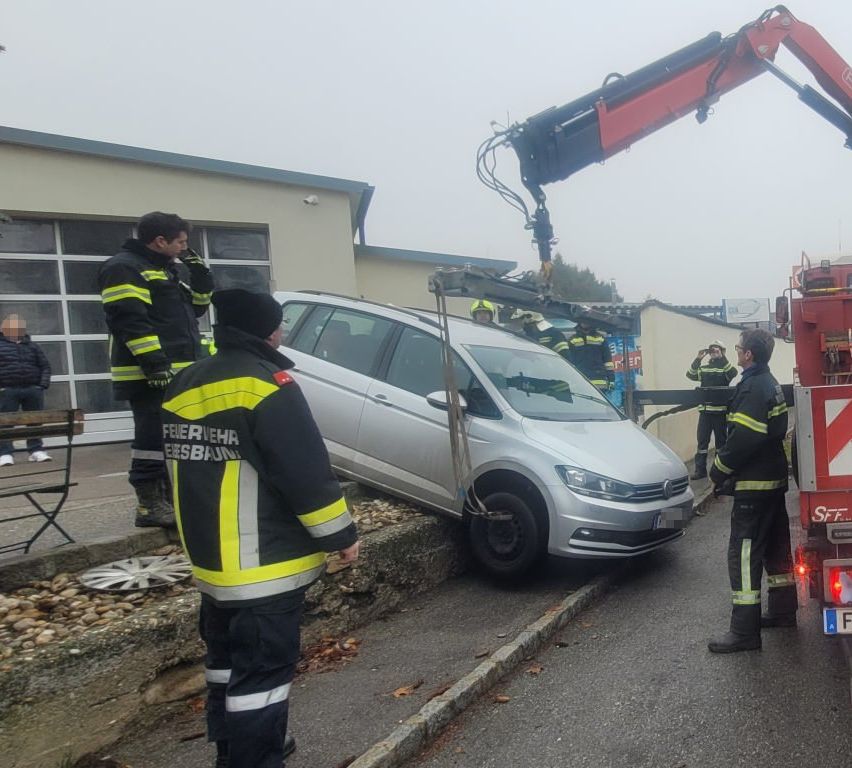 Feuerwehrleute in gelben und schwarzen Uniformen heben ein silbernes Auto mit einem Kran. Eine Person macht Fotos, während eine andere beobachtet.