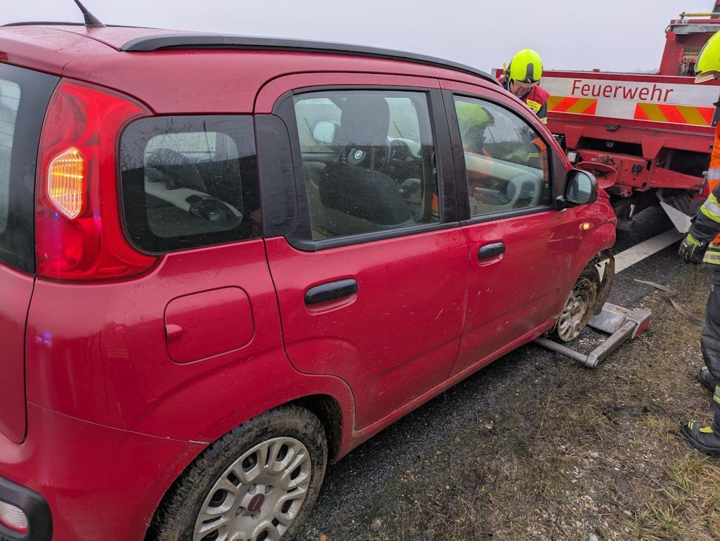 Ein rotes Auto steht am Straßenrand mit Rettungskräften in der Nähe. Das Auto hat einen Platten und ein Feuerwehrmann schneidet den Reifen mit einem Werkzeug.