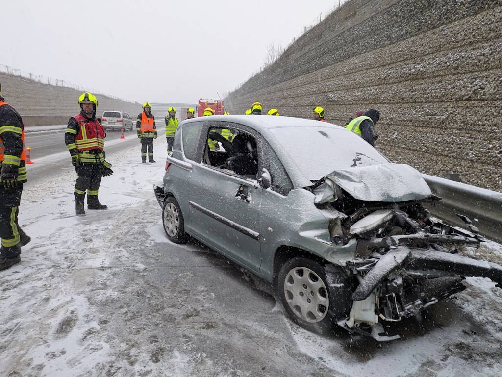 Ein demoliertes Auto steht am Straßenrand einer verschneiten Straße, während Einsatzkräfte in gelben Westen beobachten.