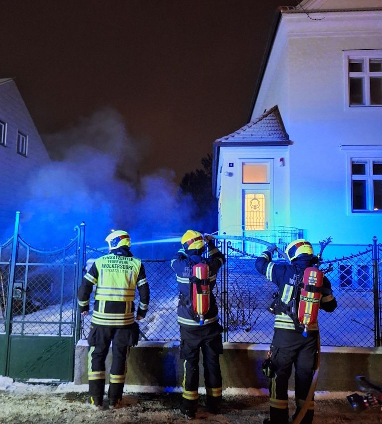 Drei Feuerwehrleute in gelben und schwarzen Uniformen sprühen Wasser mit einem Schlauch. Sie sind nachts vor einem Haus.