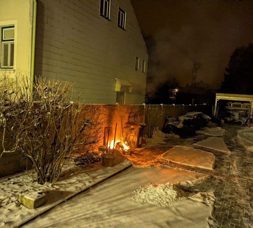 Ein kleines Feuer brennt in einem verschneiten Hinterhof. Im Hintergrund befinden sich Pflanzen, ein Holzzaun und eine Ziegelmauer.