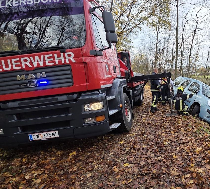 Ein roter Feuerwehrwagen ist auf einem Weg mit gefallenen Blättern geparkt. Feuerwehrleute arbeiten an einem beschädigten Fahrzeug, einer trägt einen gelben Helm. Ein Mann in einer reflektierenden Weste steht in der Nähe.