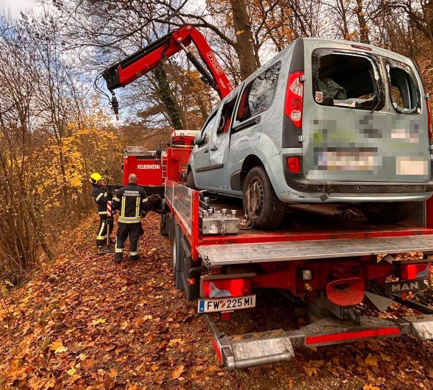 Ein roter Kran hebt einen grauen Van mit zerbrochenen Fenstern auf einen roten Flachbett-Lkw. Zwei Feuerwehrleute in gelben Helmen stehen in der Nähe. Das Bild ist unscharf.