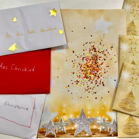 Eine Sammlung von Weihnachtskarten und Briefen auf einem Tisch mit Sternen und Glitzer. Eine weiße Karte mit goldenen Sternen trägt die Aufschrift 'An das liebe Chrisskind'. Eine rote Karte sagt 'Ans Chrisskind'. Ein weißes Papier mit einer roten Linie trägt die Aufschrift 'Chrisskind'.