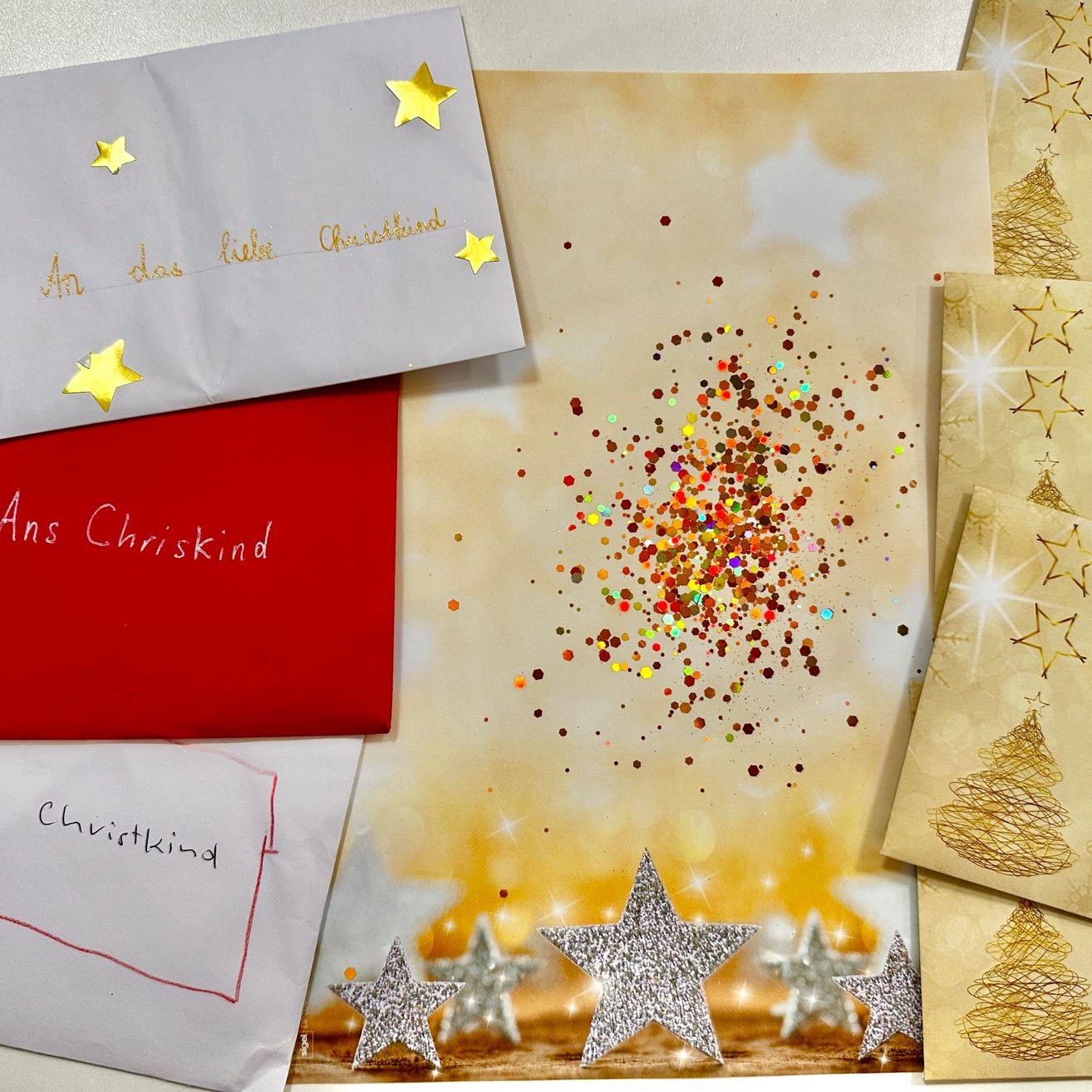 Eine Sammlung von Weihnachtskarten und Briefen auf einem Tisch mit Sternen und Glitzer. Eine weiße Karte mit goldenen Sternen trägt die Aufschrift 'An das liebe Chrisskind'. Eine rote Karte sagt 'Ans Chrisskind'. Ein weißes Papier mit einer roten Linie trägt die Aufschrift 'Chrisskind'.