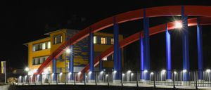 Eine Brücke mit blauen und roten Bögen und Lichtern, bei Nacht, mit einem gelben Gebäude auf der linken Seite.