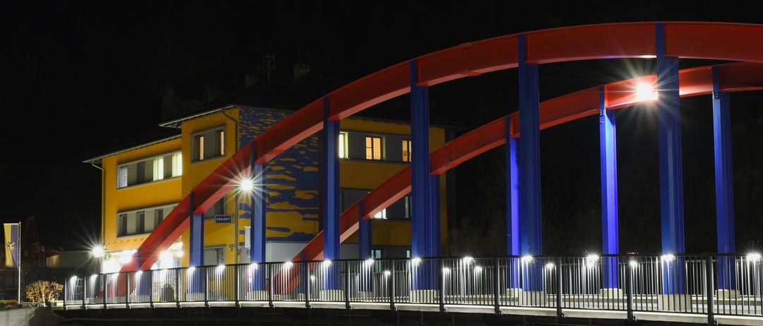 Eine Brücke mit blauen und roten Bögen und Lichtern, bei Nacht, mit einem gelben Gebäude auf der linken Seite.