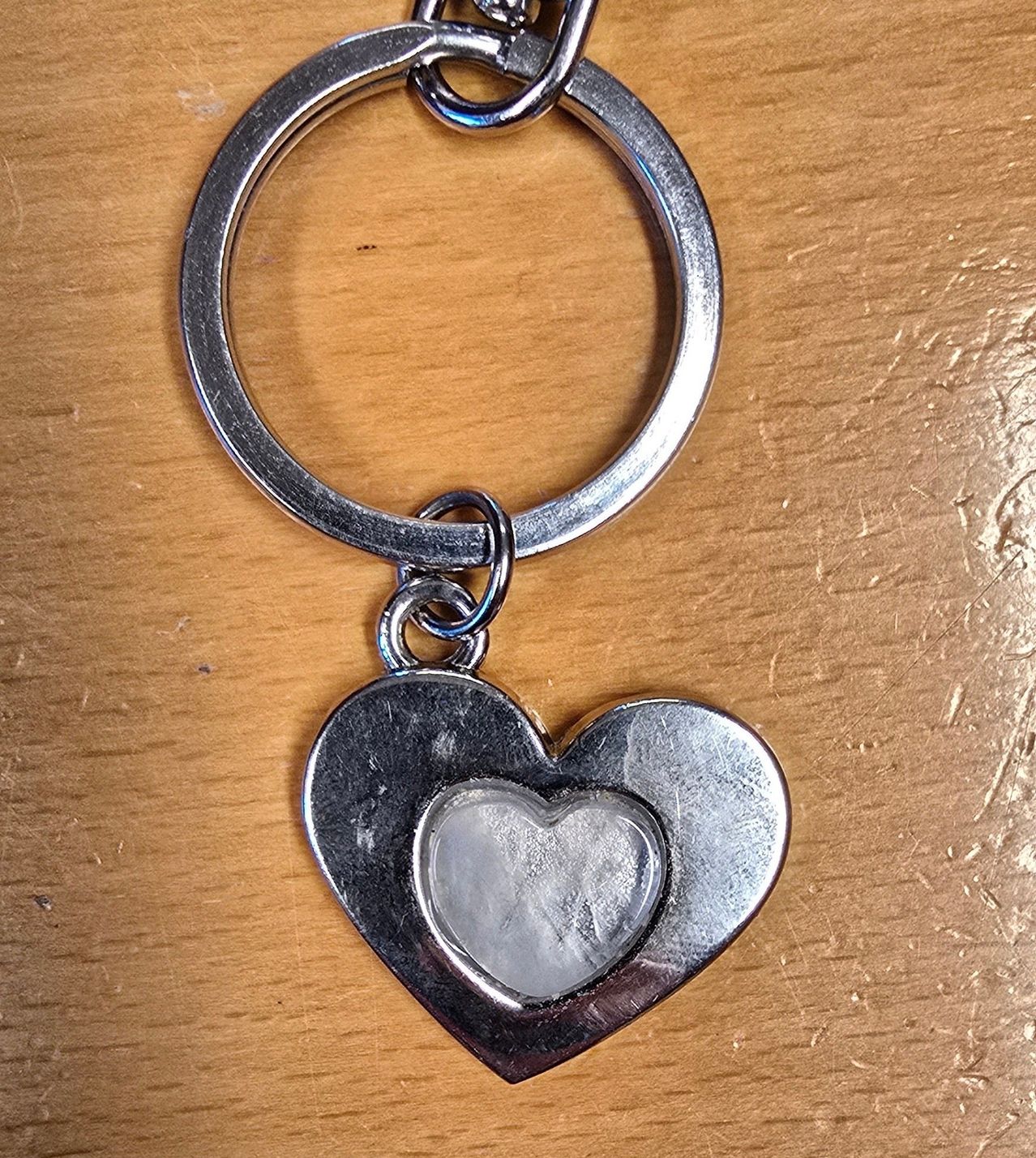 Ein silberner herzförmiger Schlüsselanhänger mit einer kleineren herzförmigen Aussparung liegt auf einer Holzoberfläche. Der Schlüsselanhänger ist an einem silbernen Ring befestigt.