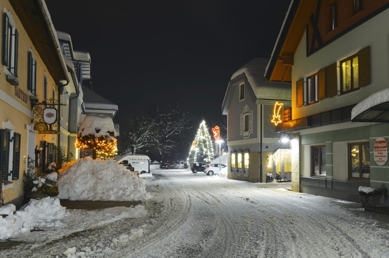 Eine verschneite Straße bei Nacht mit einem geschmückten Weihnachtsbaum, einem beleuchteten Haus und geparkten Autos. Schneehaufen liegen an den Seiten.