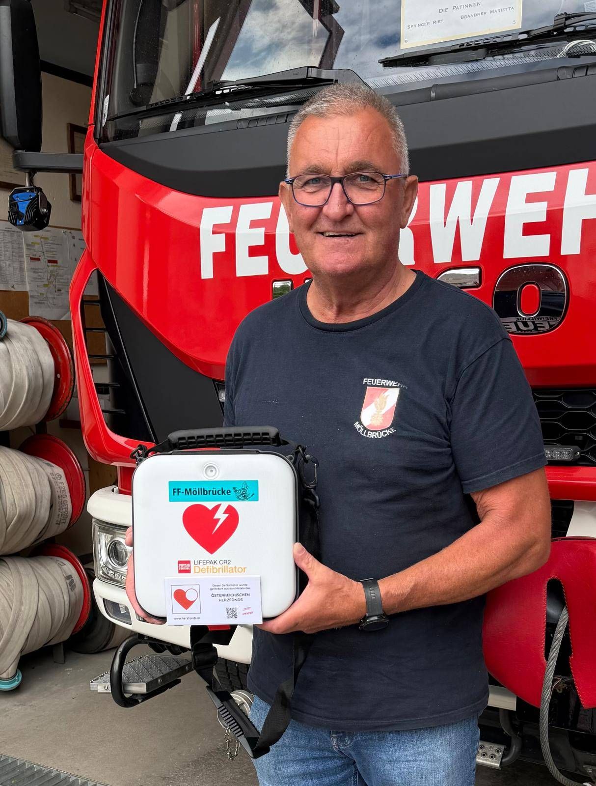 Ein Mann hält einen Defibrillator vor einem roten Feuerwehrauto. Er trägt ein marineblaues Hemd und eine Brille.