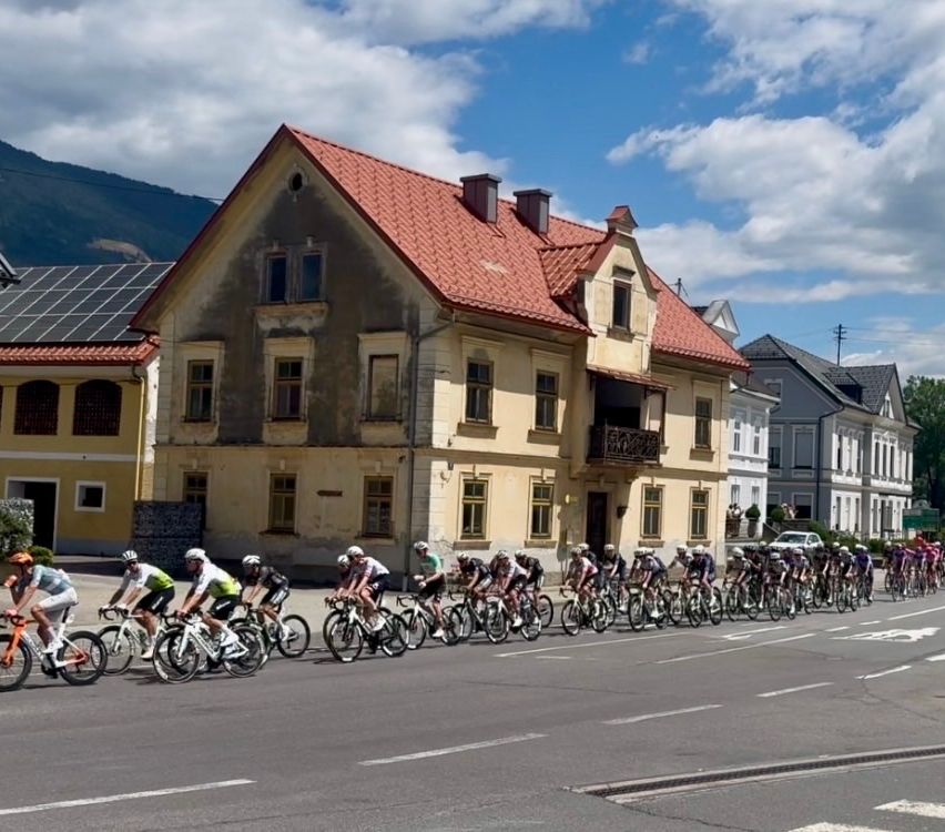Eine Gruppe von Radfahrern in Uniform fährt auf der Straße in einer Reihe mit Fahrrädern vor einem Gebäude mit rotem Dach.