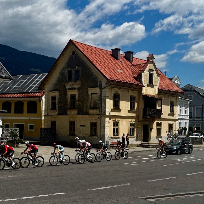 Radfahrer in Helmen fahren in einer Reihe auf einer Stadtstraße, mit Gebäuden und Bergen im Hintergrund.