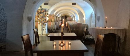 A candlelit room with wooden tables and chairs, an arched ceiling, and wine barrels. The room is dimly lit, with a table covered by a white cloth.